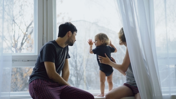 Happy family with young cute daughter sitting on windowsill playing and looking in window at home in the morning